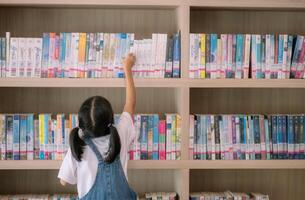 uma jovem menina é alcançando para uma livro em uma biblioteca estante foto