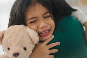 uma jovem menina é abraçando uma Urso de pelúcia Urso enquanto chorando foto