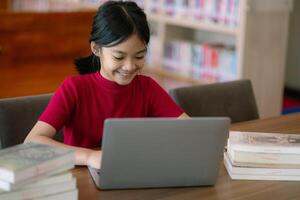 uma jovem menina é sentado às uma mesa com uma computador portátil e uma pilha do livros foto