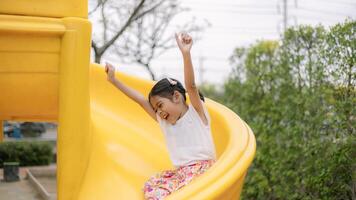 uma jovem menina é sentado em uma amarelo deslizar, sorridente e acenando foto