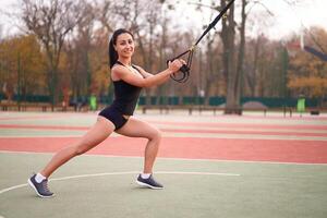 menina atleta Treinamento usando trx em campo esportivo. misturado raça jovem adulto mulher Faz exercite-se com suspensão sistema. saudável estilo de vida. alongamento ao ar livre Parque infantil. foto