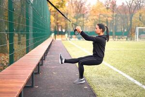 menina atleta Treinamento usando trx em campo esportivo. misturado raça jovem adulto mulher Faz exercite-se com suspensão sistema. saudável estilo de vida. alongamento ao ar livre Parque infantil. foto