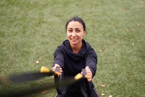 menina atleta funcional Treinamento em campo esportivo. misturado raça jovem adulto mulher Faz exercite-se com suspensão sistema. saudável estilo de vida. alongamento ao ar livre Parque infantil. foto