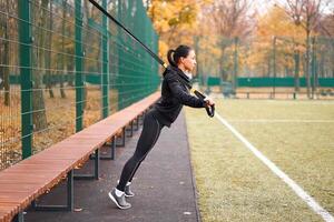 menina atleta Treinamento usando trx em campo esportivo. misturado raça jovem adulto mulher Faz exercite-se com suspensão sistema. saudável estilo de vida. alongamento ao ar livre Parque infantil. foto