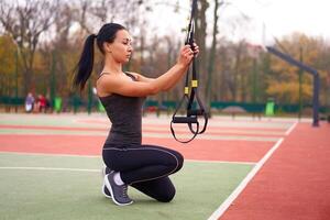 menina atleta Treinamento usando trx em campo esportivo. misturado raça jovem adulto mulher Faz exercite-se com suspensão sistema. saudável estilo de vida. alongamento ao ar livre Parque infantil. foto