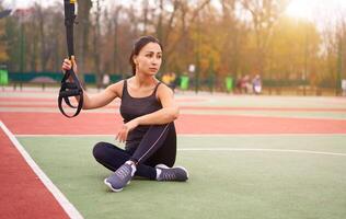 menina atleta funcional Treinamento em campo esportivo. misturado raça jovem adulto mulher Faz exercite-se com suspensão sistema. saudável estilo de vida. alongamento ao ar livre Parque infantil. foto