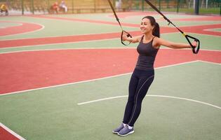 menina atleta Treinamento usando trx em campo esportivo. misturado raça jovem adulto mulher Faz exercite-se com suspensão sistema. saudável estilo de vida. alongamento ao ar livre Parque infantil. foto