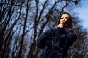 ao ar livre foto do uma glamour mulher com Sombrio cabelo dentro pele casaco em pé cruzado mãos, posando dentro inverno parque