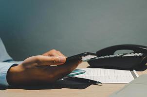 mão segurando o telefone inteligente na mesa com papelada do documento e telefone no escritório. foto