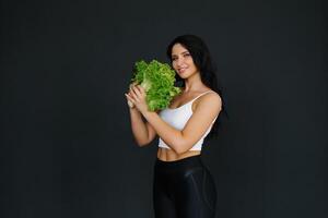 jovem fêmea ginástica treinador ou nutricionista segurando alface folhas em uma Preto fundo e sorridente foto