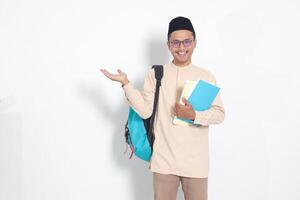 retrato do animado aluna ásia muçulmano homem dentro Koko camisa com calota craniana carregando mochila, segurando escola livros, apontando para a lado. islâmico Educação conceito. isolado imagem em branco fundo foto