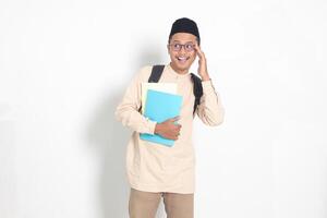 retrato do animado aluna ásia muçulmano homem dentro Koko camisa com calota craniana carregando mochila, enquanto segurando dele escola livros. islâmico Educação conceito. isolado imagem em branco fundo foto