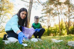 mulher voluntário e pequeno Garoto colheita acima a plástico lixo e colocando isto dentro biodegradável saco do lixo ao ar livre. ecologia, reciclando e proteção do natureza conceito. de Meio Ambiente proteção. foto