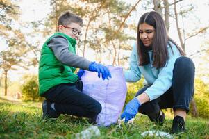 mulher voluntário e pequeno Garoto colheita acima a plástico lixo e colocando isto dentro biodegradável saco do lixo ao ar livre. ecologia, reciclando e proteção do natureza conceito. de Meio Ambiente proteção. foto