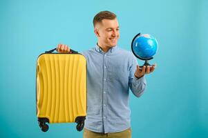 alegre viajante turista homem dentro casual roupas com mala de viagem isolado em azul fundo. masculino passageiro viajando no exterior em fim de semana. ar voar viagem conceito aguarde mundo globo foto