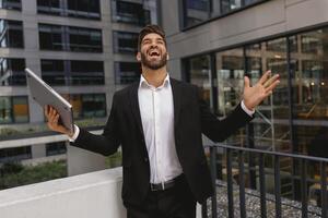 homem de negocios com computador portátil em pé em escritório terraço e elevado mãos é satisfeito do acabado trabalhos foto