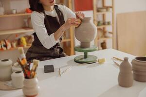 fechar acima do jovem fêmea ceramista vestindo avental trabalhos com não disparado argila vaso dentro cerâmica estúdio foto