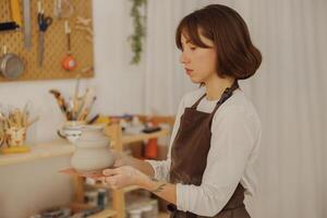 jovem fêmea oleiro segurando não disparado argila vaso enquanto em pé dentro cerâmica estúdio. Alto qualidade foto