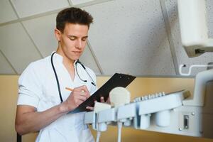 retrato do uma bonito jovem médico Próximo para a ultrassom digitalização máquina. ele é apertando os olhos, olhando às a Câmera com desconfiado olhar, o preenchimento pacientes história. foto