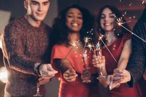 sorrindo e se sentindo feliz. amigos multirraciais celebram o ano novo segurando luzes de bengala e copos com bebida