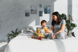 mãe com filho dentro pijamas comendo fruta em cama. retrato feliz, sorridente, alegre família, mãe e filho tendo café da manhã dentro cama, surpresa em mãe dia. positivo humano emoções foto