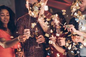 confete no ar. amigos multirraciais celebram o ano novo segurando luzes de bengala e copos com bebida