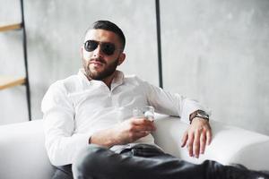 vista frontal. foto de jovem empresário barbudo com óculos escuros e uísque na mão, sente-se no sofá branco
