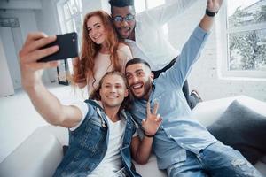 rosto fazendo careta. jovens amigos alegres tirando selfies no sofá e no interior branco foto