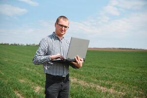 agricultor em pé dentro uma trigo campo e olhando às computador portátil, estão examinando corporação. jovem bonito agrônomo. agronegócio conceito. agrícola engenheiro em pé dentro uma trigo campo foto