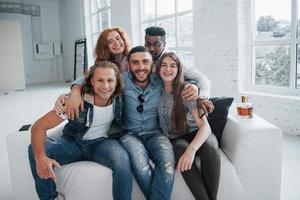 jovens amigos engraçados se divertindo sentados no sofá no interior branco foto