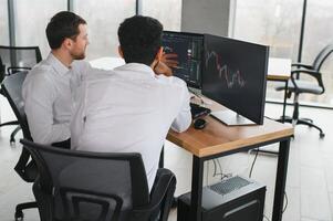 dois homens comerciantes sentado às escrivaninha às escritório juntos monitoramento ações dados vela gráficos em tela analisando preço fluxo sorridente alegre tendo lucro trabalho em equipe conceito foto