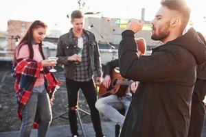 cara elegante bebe cerveja. festa no telhado. amigos com álcool se divertindo. cara tocando violão foto