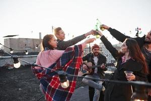 para a felicidade. grupo de jovens comemorando em um telhado com um pouco de álcool e tocando violão foto