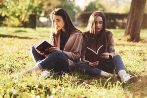 jovens irmãs gêmeas morenas sentadas perto uma da outra na grama com as pernas ligeiramente dobradas nos joelhos e cruzadas, lendo livros marrons, vestindo um casaco casual no parque ensolarado de outono em fundo desfocado foto