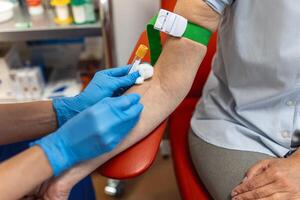 preparação para sangue teste com Senior mulher de fêmea médico médico uniforme em a mesa dentro branco brilhante sala. enfermeira pierces a pacientes braço veia com agulha em branco tubo. foto