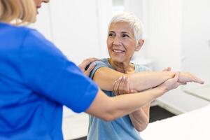 feliz Senior mulher fazendo exercício com fisioterapeuta. velho aposentado senhora fazendo alongamento braços às clínica com a Socorro do uma pessoal treinador durante uma reabilitação sessão. foto