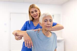 fêmea fisioterapeuta trabalhando examinando tratando ferido braço do Senior fêmea paciente, alongamento e exercício, fazendo a reabilitação terapia dor dentro clínica. foto