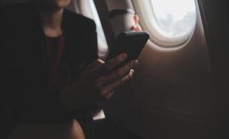 Loiras fêmea turista verificação entrada notificação em Smartphone sentado em assento do avião com netbook.jovem empresária compartilhar meios de comunicação a partir de Telefone em computador portátil computador durante avião voar foto