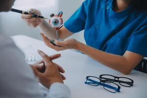 fechar-se do ásia fêmea médico falando com idosos paciente mostrando globo ocular modelo e explicando olho doença dentro hospital foto