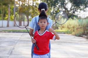 ásia Garoto tem dele costas para dele Mais velho irmã, segurando uma badminton raquete e uma branco peteca. foto
