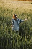 jovem bonita mulher dentro vermelho verão vestir e Palha chapéu caminhando em amarelo Fazenda campo com maduro dourado trigo desfrutando caloroso tarde. foto