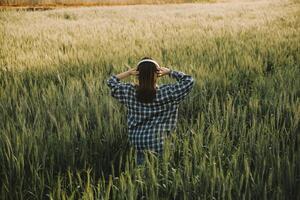 jovem bonita mulher dentro vermelho verão vestir e Palha chapéu caminhando em amarelo Fazenda campo com maduro dourado trigo desfrutando caloroso tarde. foto