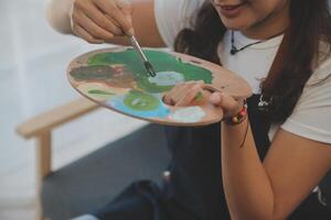 cortada imagem do fêmea artista em pé dentro frente do a cavalete e mergulho escova para dentro cor paleta foto