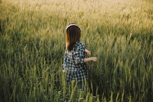 jovem bonita mulher dentro vermelho verão vestir e Palha chapéu caminhando em amarelo Fazenda campo com maduro dourado trigo desfrutando caloroso tarde. foto