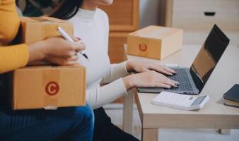 mulheres de negócios asiáticas usam laptop verificando caixas de remessa on-line de pedidos de clientes em casa. começando pequeno empresário freelance. negócio online, trabalho em casa conceito. foto