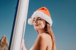 mulher mar e aí. fechar acima retrato do feliz jovem caucasiano mulher com grandes cabelo dentro santa chapéu olhando às Câmera e sorridente. fofa mulher retrato dentro uma branco bikini posando em sup borda dentro a mar foto