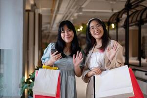 dois alegre jovem ásia mulheres onda alegremente às a Câmera enquanto segurando compras bolsas. foto