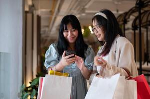 dois ásia mulheres bate-papo alegremente dentro uma compras Shopping corredor, olhando às uma Smartphone junto. foto