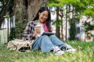uma jovem ásia mulher senta debaixo uma árvore dentro uma campus parque, segurando uma café copo e uma tábua. foto