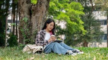 uma jovem ásia mulher senta debaixo uma árvore dentro uma campus parque, segurando uma café copo e uma tábua. foto